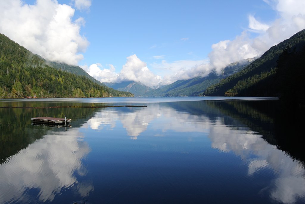 A calm lake—one of the best boating lakes in Washington State—reflects clouds and forested mountains under a blue sky, with a small dock floating near the shore.
