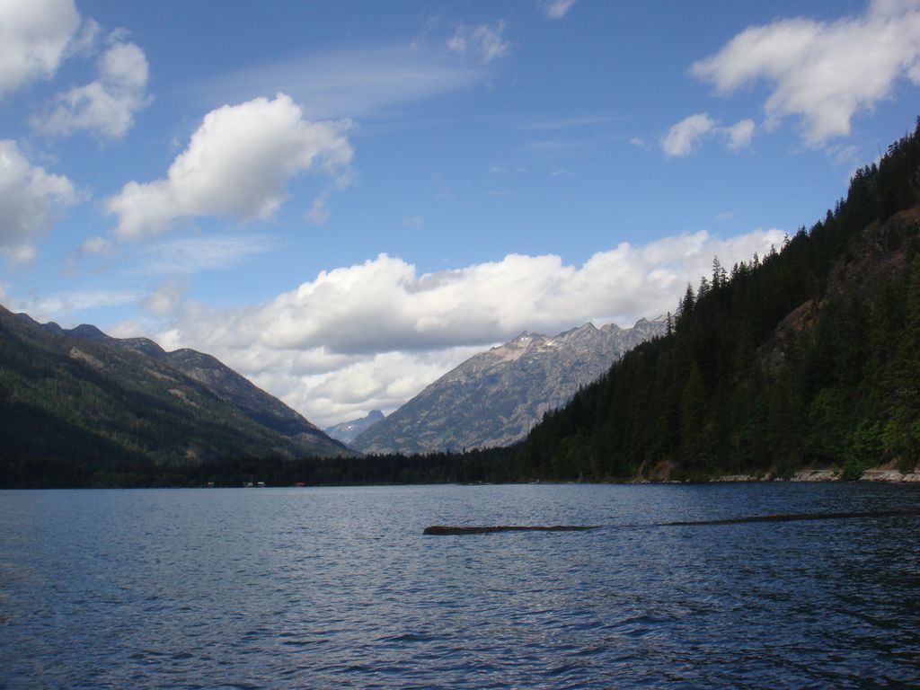 A calm lake, ranked among the top boating lakes in Washington State,'s surrounded by forested hills and far-off snow-capped mountains beneath a partly cloudy sky.