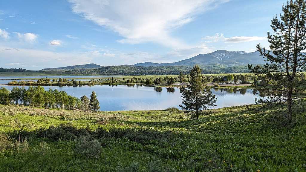 Lake, one of the best boating lakes in Colorado, is surrounded by green grass, scattered trees, and distant mountains under a partly cloudy blue sky.