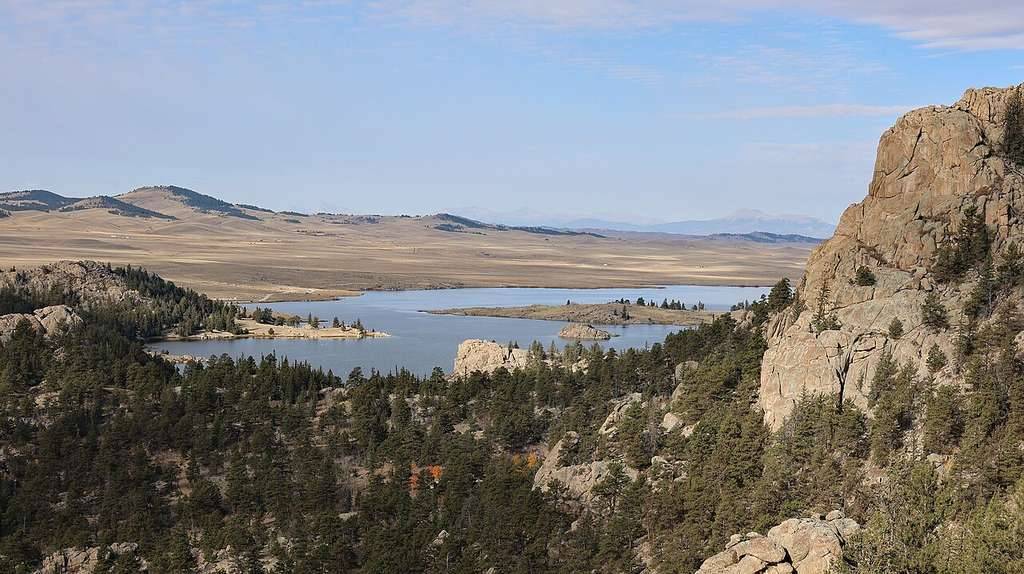 Rocky cliffs frame a winding river—one of Colorado's best boating lakes—surrounded by Evergreens, open plains, and distant hills under a partly cloudy sky.