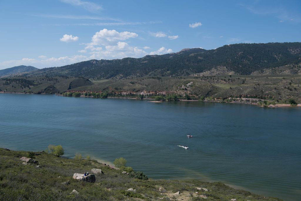 A wide Colorado lake—one of the best for boating—has two small boats, surrounded by green hills and mountains under a partly cloudy sky. No visible fish like Trout or Bass in sight.