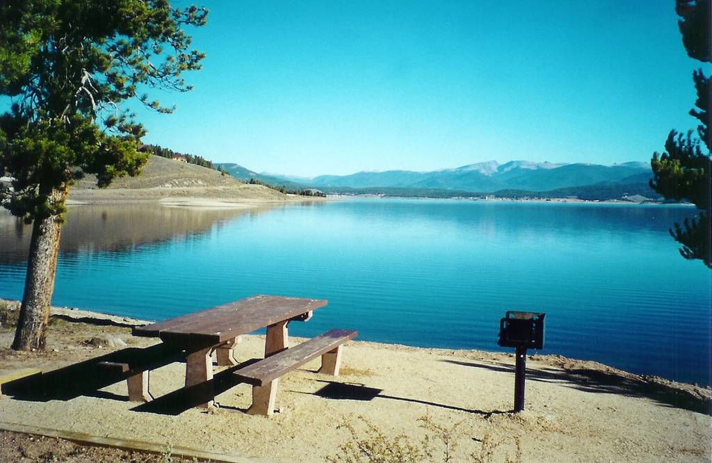 There's a wooden picnic table and grill on sandy ground by a calm lake—one of Colorado's top boating lakes. Mountains rise behind, and the sky's clear blue. No fish or species names are mentioned.