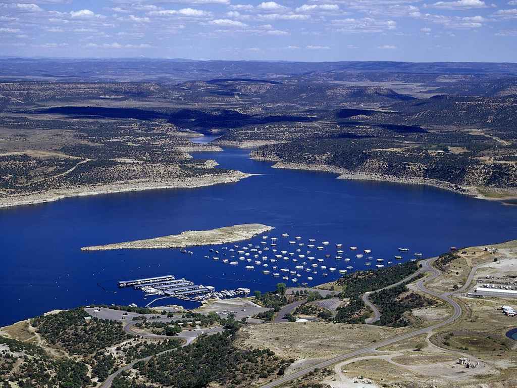 Aerial view of one of Colorado's best boating lakes with marinas, boats, parking lots, roads, and dry, hilly land under a partly cloudy sky.