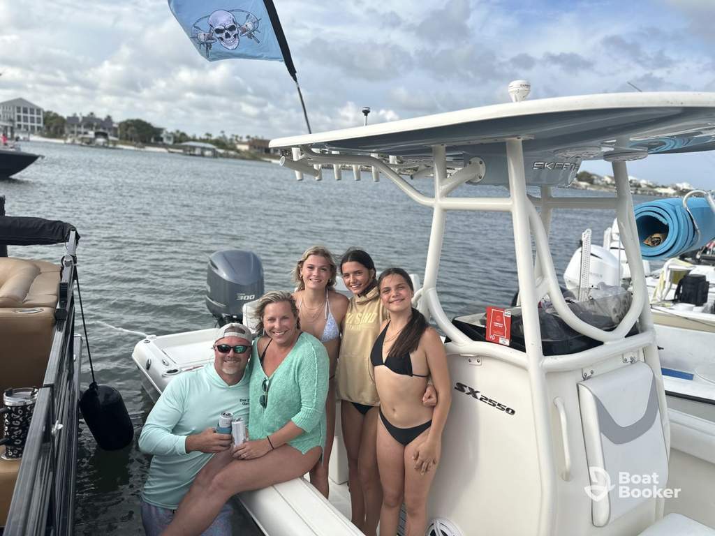 Five people pose on a docked white boat—four women stand, and one man with an Alabama boating license sits holding a drink. Water, boats, and houses are under a partly cloudy sky.