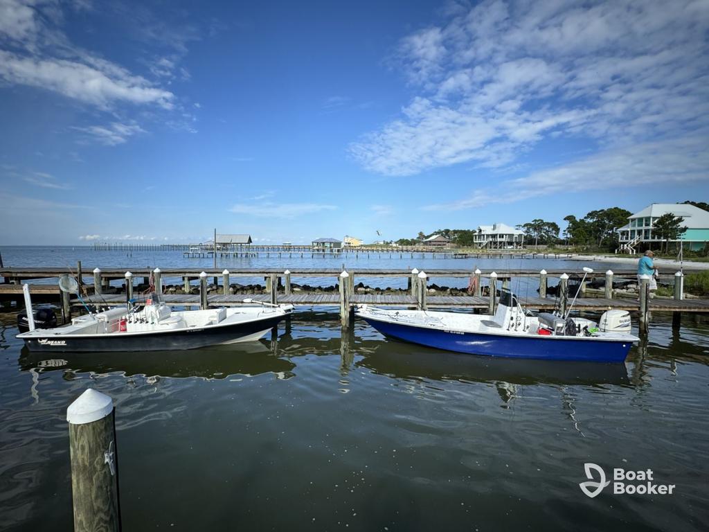 Two small motorboats are docked at a wooden pier—great if you've got an Alabama boating license—with houses and a long pier behind them under a partly cloudy blue sky.