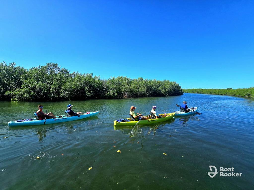 Four people in two kayaks're paddling along a calm, tree-lined river under a clear blue sky. The "Boat Booker" logo's in the lower right corner. No fish or species' names appear here.