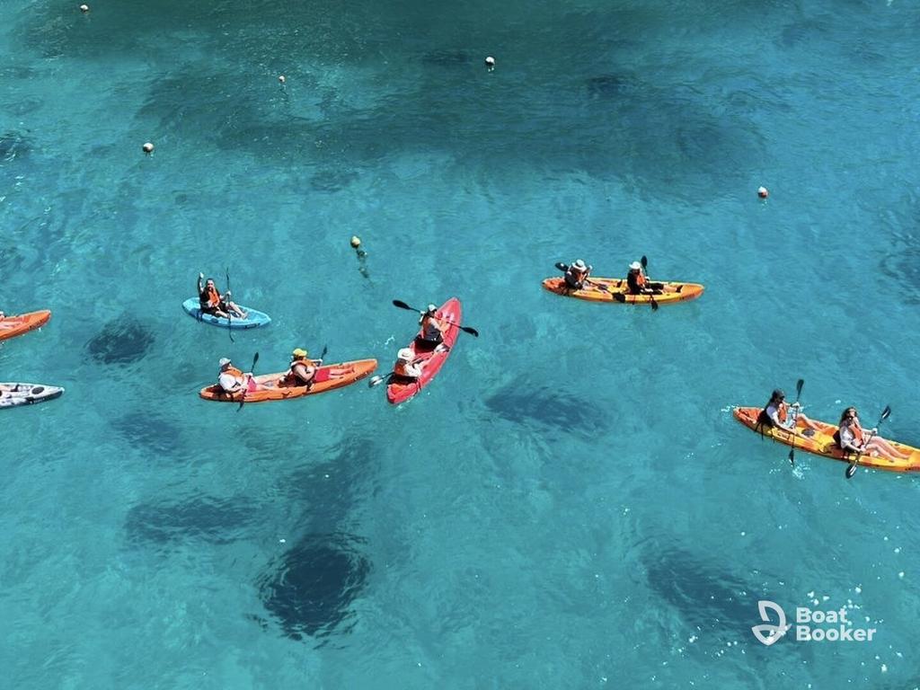 Seven kayaks with people paddling float on clear blue water. You can see their shadows below. The "Boat Booker" logo's in the lower right corner.