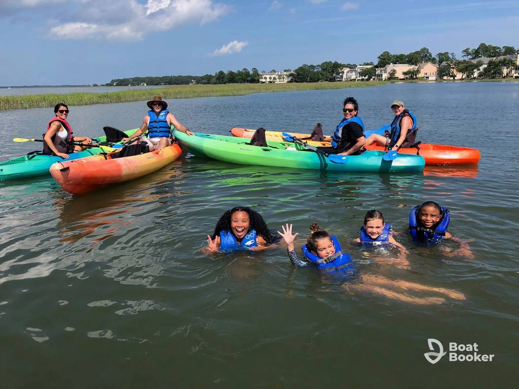 Four kids in blue life jackets swim and wave at the camera near three kayaks with five people on a calm lake, with houses and trees behind them.