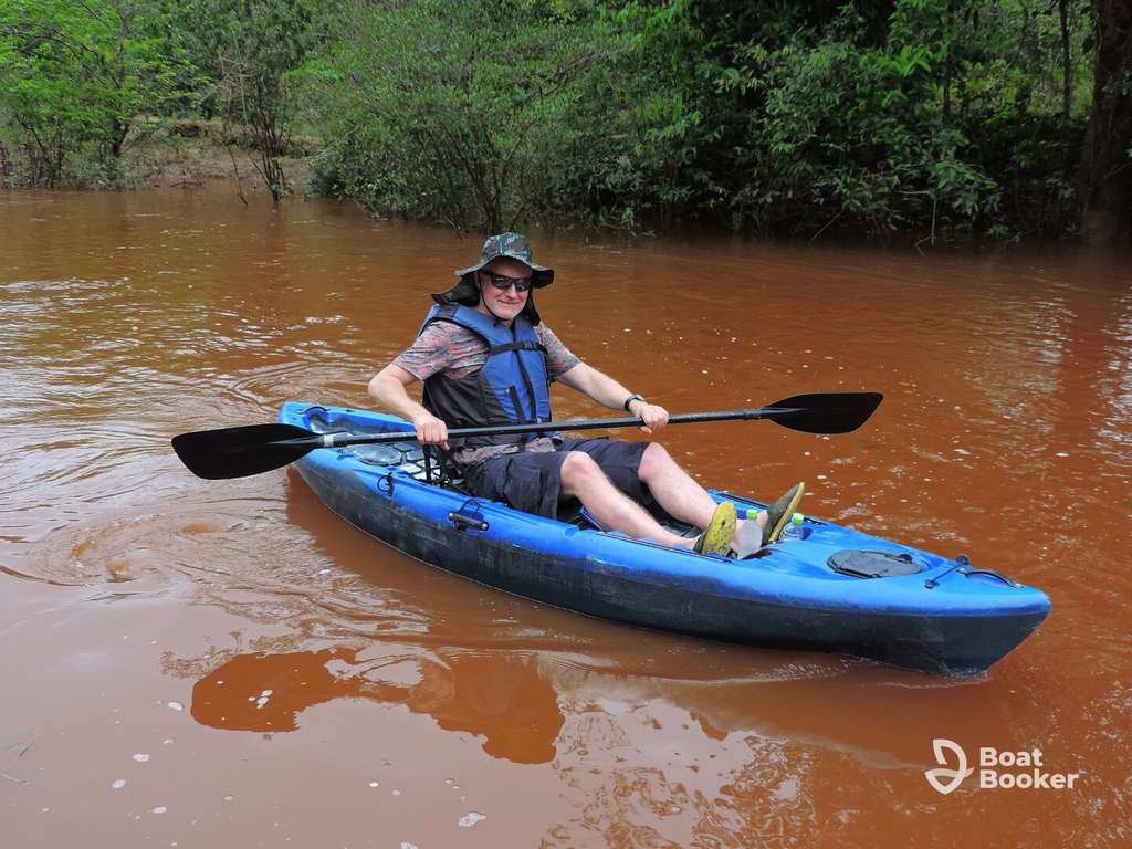 Someone's wearing a hat and life jacket while paddling a blue kayak on a muddy river, with green trees all around. The "Boat Booker" logo's in the bottom right corner.