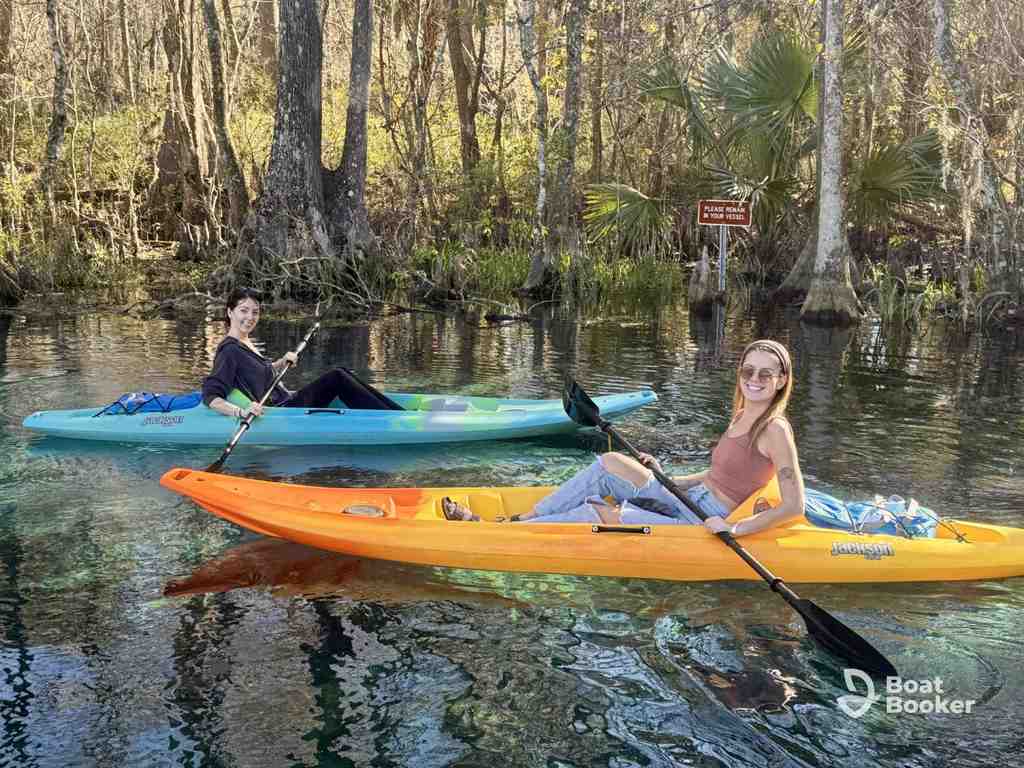 Two people in kayaks paddle on a clear, calm river surrounded by trees and vegetation. There's a small sign in the background.