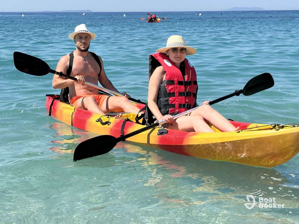 Two people wearing hats sit in a yellow and red kayak on clear blue water, holding paddles. One person's got a life jacket on. No fish like Trout or Salmon are visible in the image.