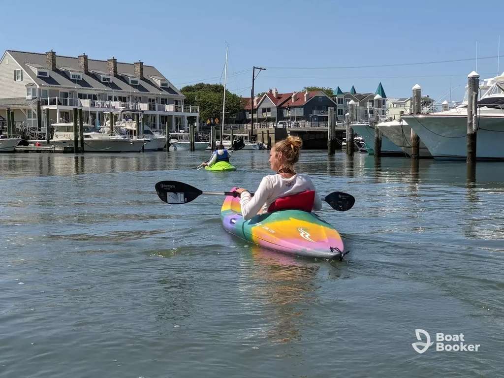 Two people are kayaking on calm water near a marina with boats and houses in the background under a clear sky.