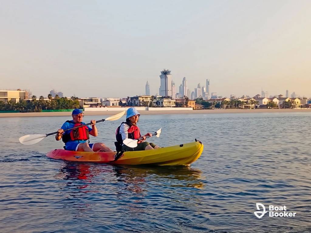 Two people are kayaking on calm water by a city shoreline under a clear sky, with buildings in the background and the Boat Booker logo in the corner.