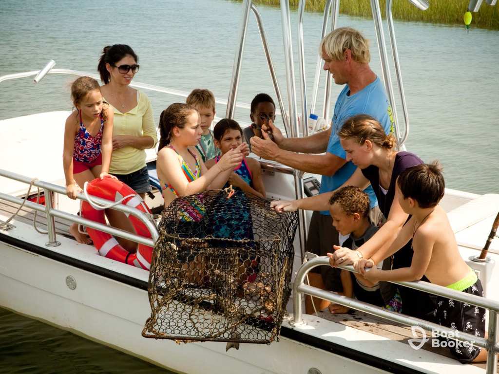 A group of adults and children on a boat watch as a guide holds up a wire mesh trap with a Crab inside.
