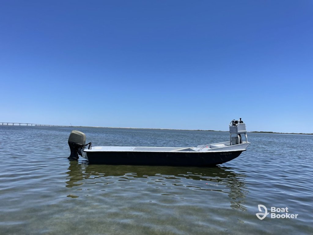 A small skiff floats on calm, shallow water under a clear blue sky. A distant bridge and land line the horizon. The Boat Booker logo's in the bottom right corner.