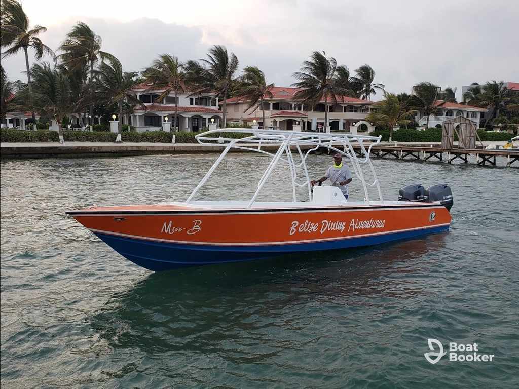 A man’s driving an orange and blue skiff named "Miss B Belize Diving Adventures" on the water, with waterfront villas and palm trees behind him.