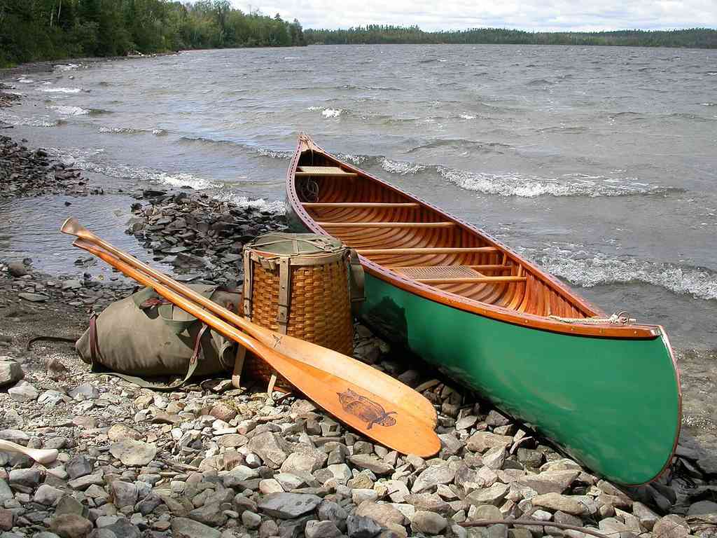 There's a green canoe on a rocky lakeshore next to a pack basket, a duffel bag, and a wooden paddle. Trees line the far shore beneath cloudy skies.