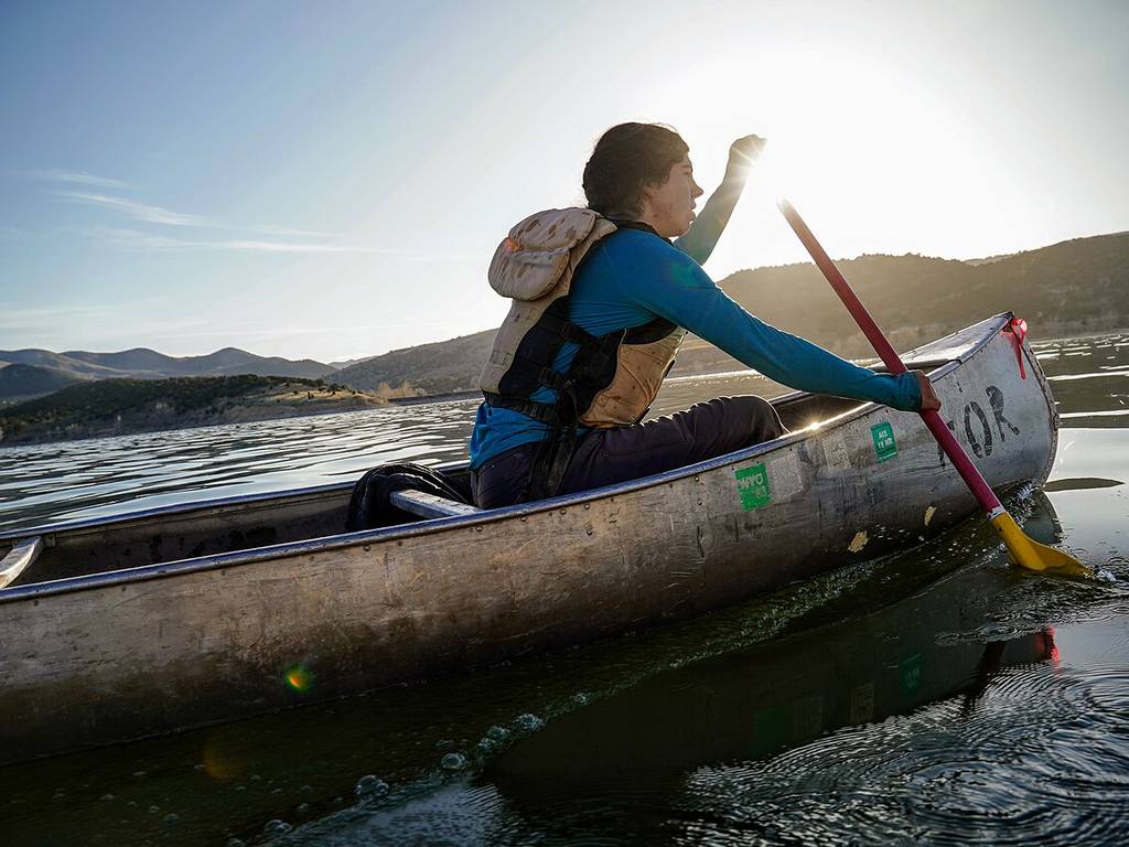 Someone's paddling a canoe on a lake at sunrise or sunset, wearing a life vest. There are hills in the background and sunlight's reflecting on the water.