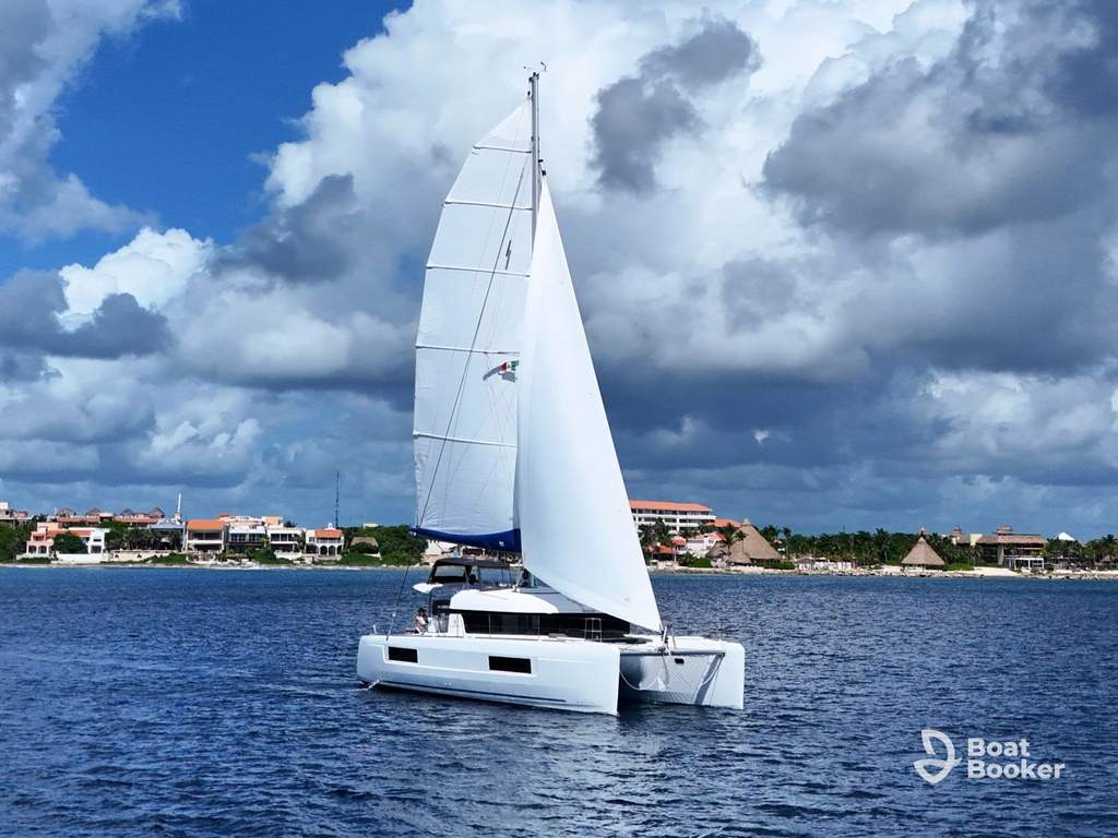 A white catamaran, great for Sailing 101, glides on blue water near a shoreline with buildings and greenery under a partly cloudy sky.