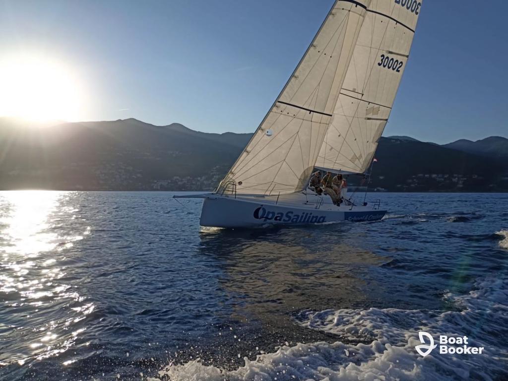 A white sailboat with “Opa Sailing” on its side glides through calm water by mountains at sunset, showing sailing’s peaceful spirit—sailing one-o-one at its best.