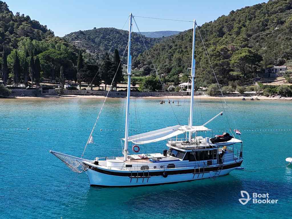 A white sailboat with two masts floats on clear blue water by a forested shore, giving off a Sailing 101 vibe; the “Boat Booker” logo’s in the corner.