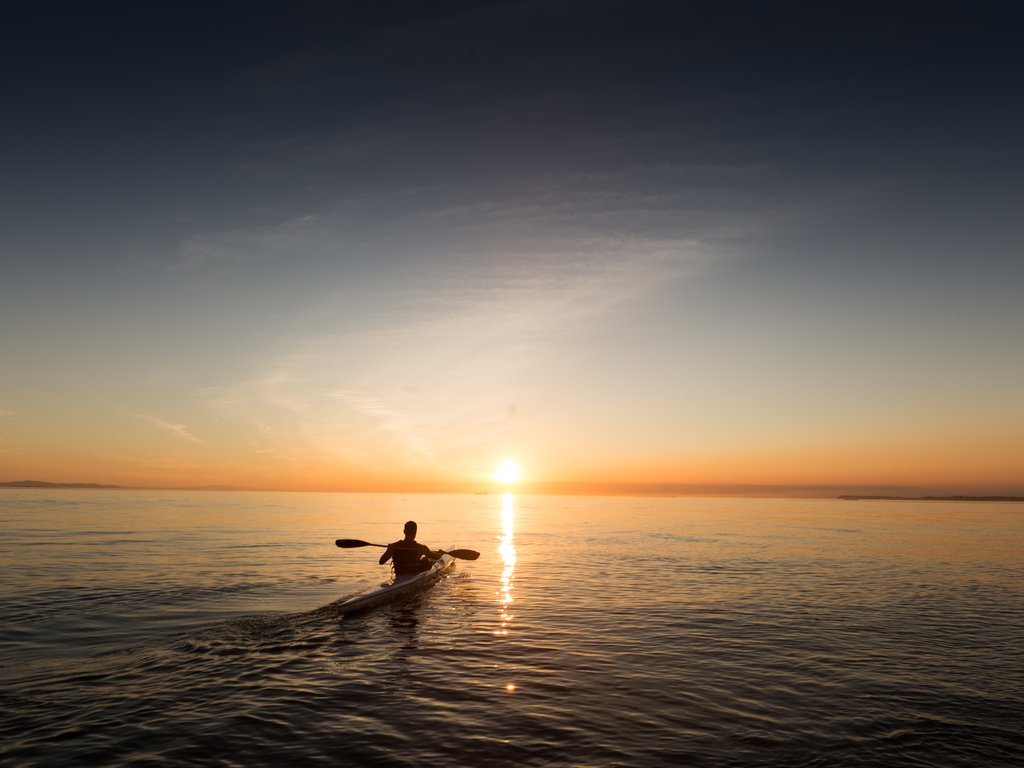 Someone’s showing how to kayak on calm water during a vibrant sunset, with the sun low near the horizon and a clear sky overhead.