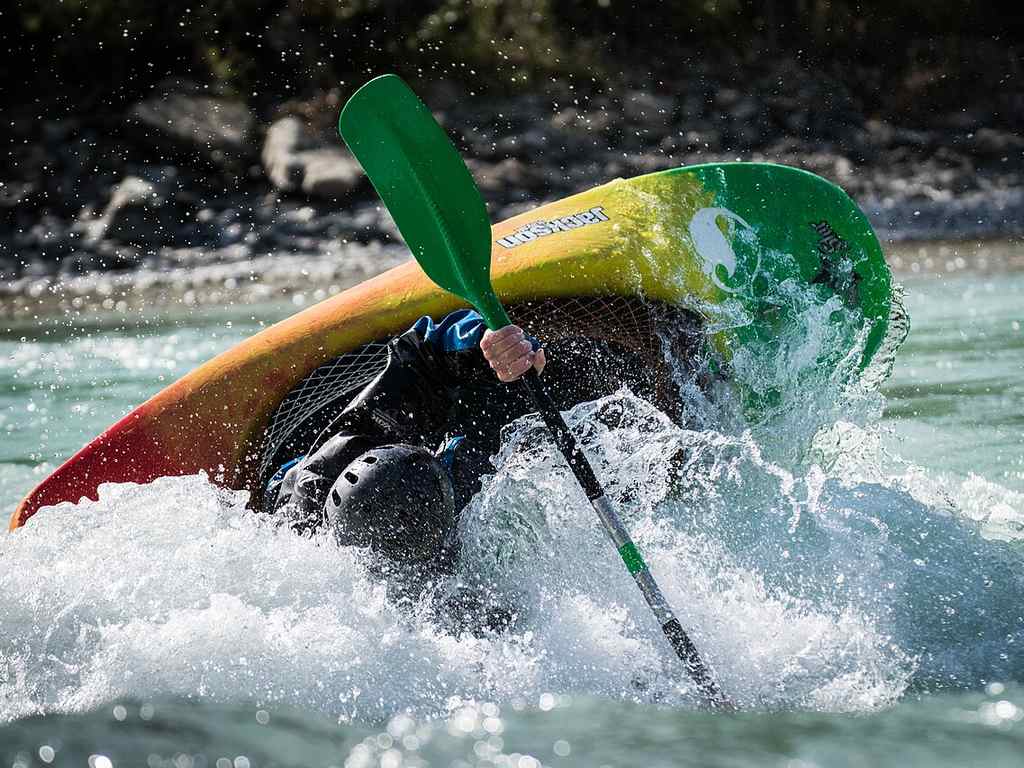 A kayaker’s wearing a helmet, showing how to kayak through rough river water as the kayak tilts and water splashes all around. No fish like Trout or Salmon are visible in the scene.