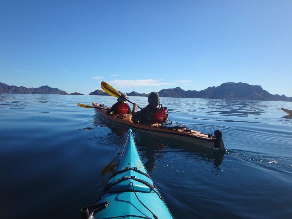 Two people show how to kayak in a double kayak on calm water, surrounded by mountains and a clear blue sky, as seen from another kayak.