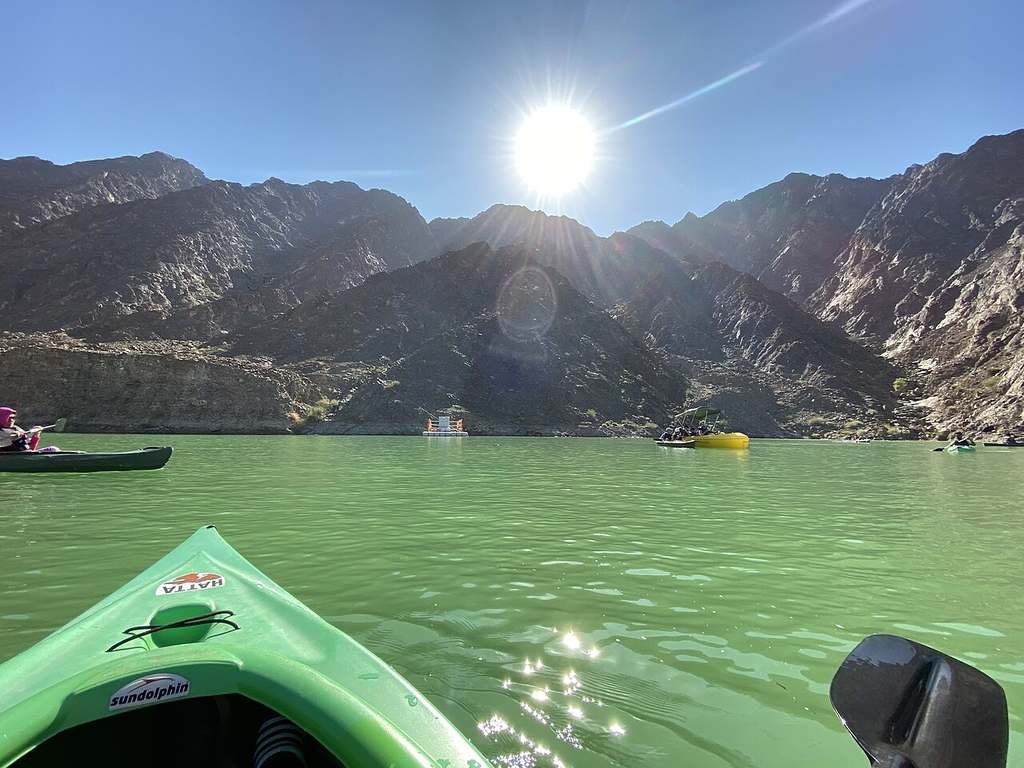 A green kayak’s floating on calm, greenish water with others around, rocky mountains and bright sun behind—it’s perfect for anyone learning how to kayak.