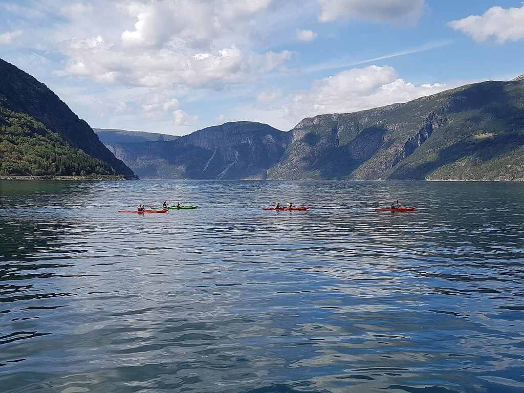 Four kayaks with people paddle on a calm lake as they’re learning how to kayak, surrounded by mountains under a partly cloudy sky.