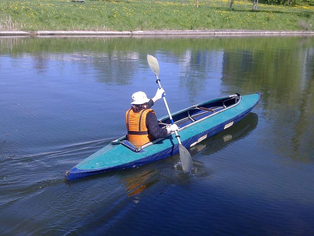 Someone’s wearing an orange life jacket and white hat, showing how to kayak as they paddle a blue kayak on calm water by a grassy shore.