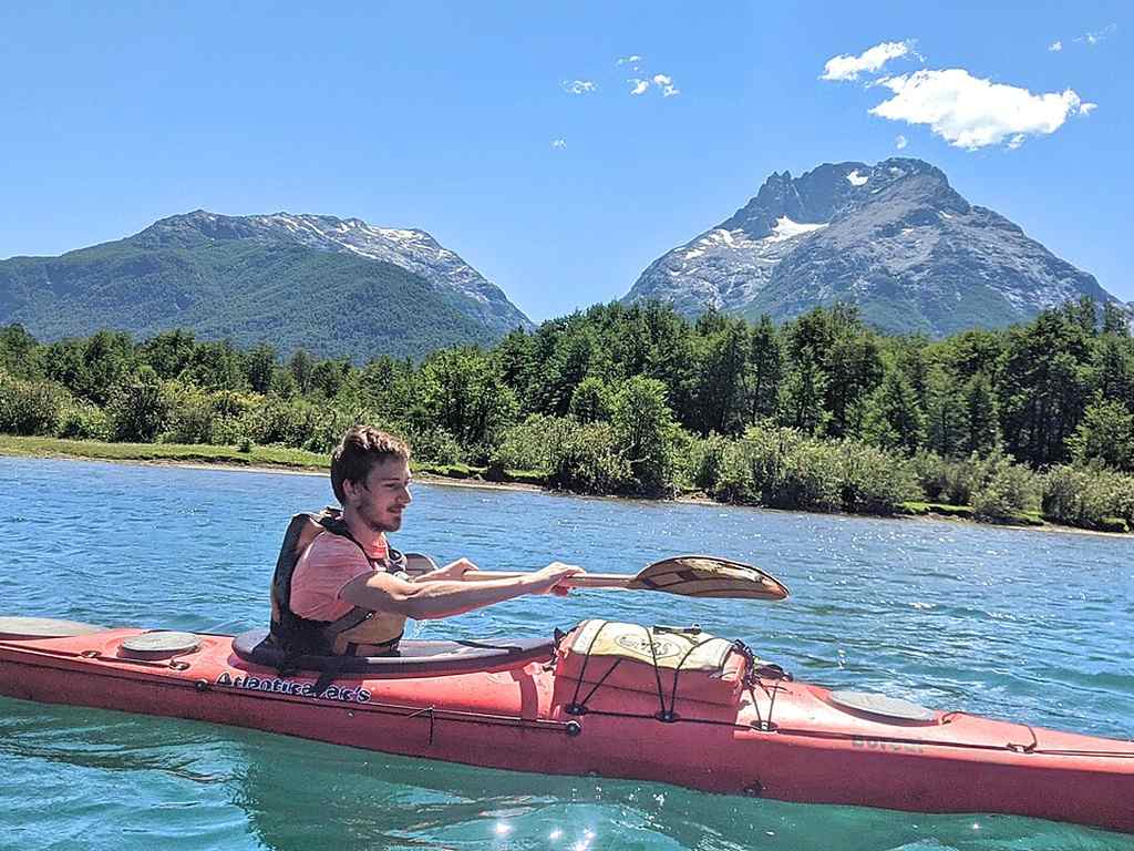 Someone’s showing how to kayak, paddling a red kayak on a lake surrounded by green trees and two snow-capped mountains under a clear blue sky.