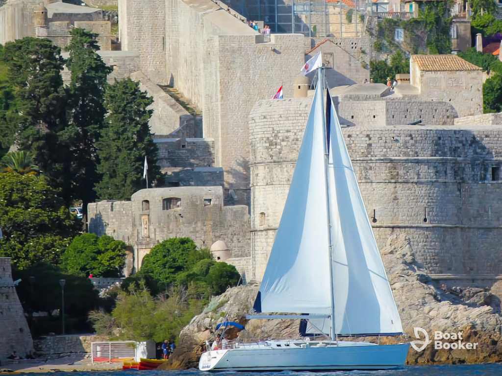 A white sailboat’s gliding by stone fortress walls and lush greenery on a sunny day, capturing Croatia’s sailing vibe; “Boat Booker” logo’s in the bottom right corner.