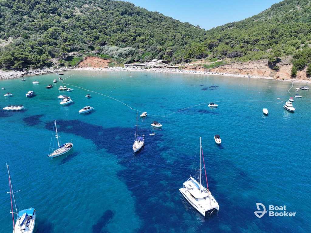 An aerial view shows a bay’s clear blue water with several sailboats and yachts anchored—ideal for sailing in Croatia—set against a tree-covered hillside in the background.