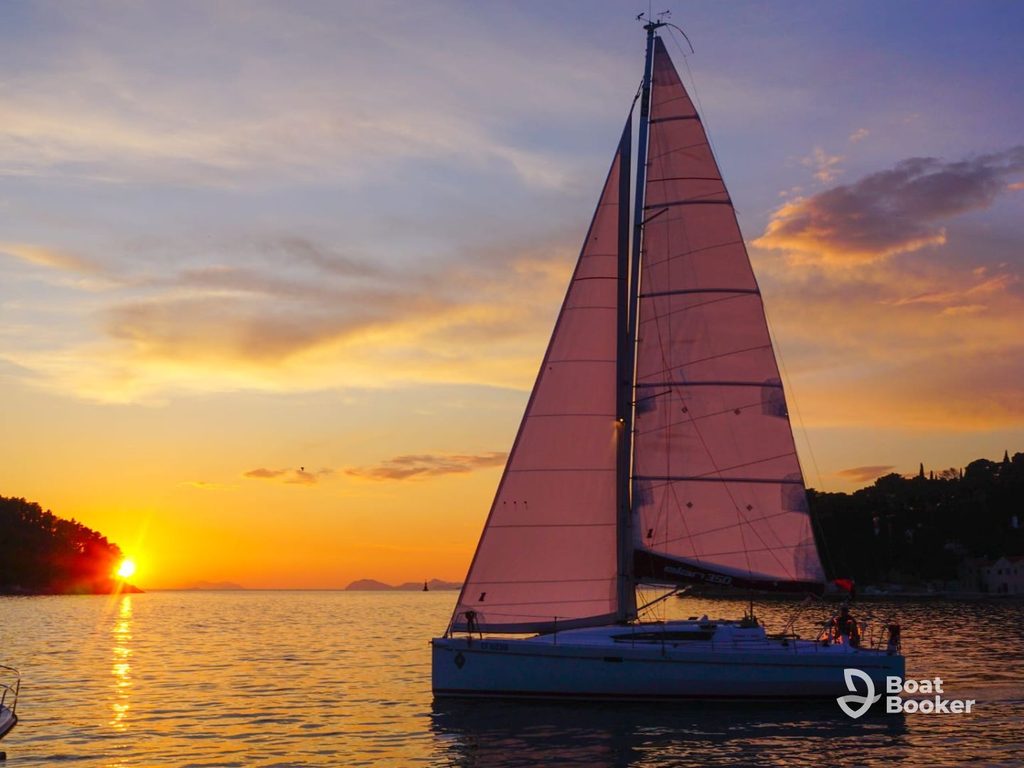 A sailboat’s gliding on calm water at sunset, orange and purple hues above—ideal for sailing in Croatia. The Boat Booker logo’s in the bottom right corner.