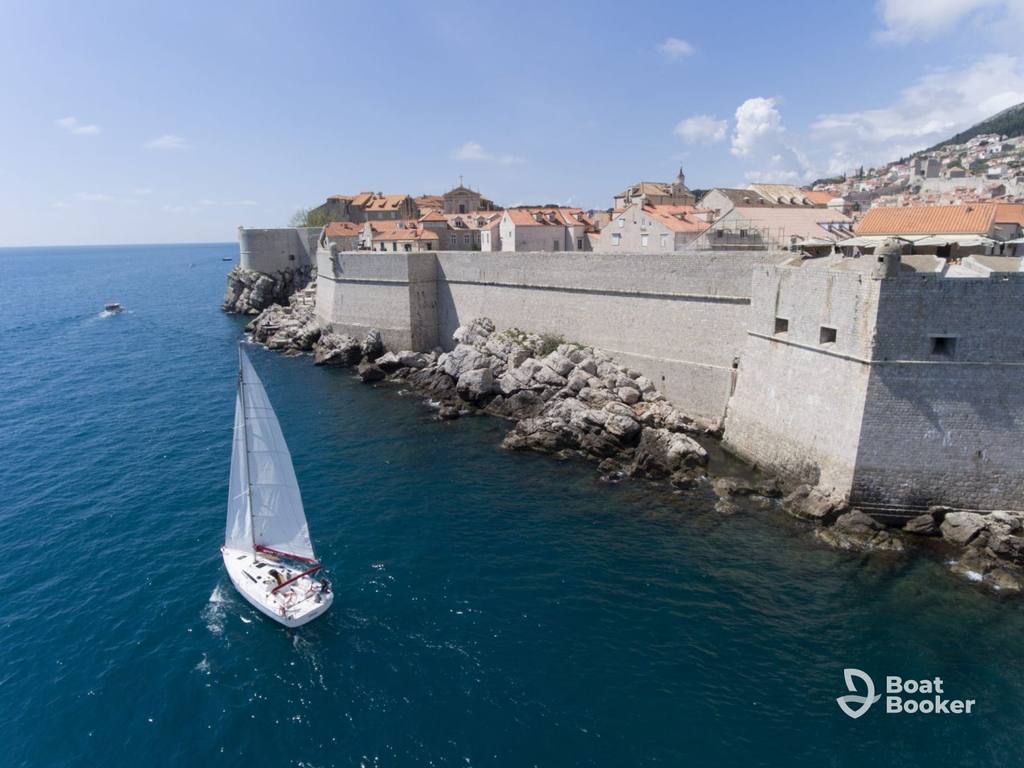 A white sailboat’s sailing in Croatia by stone city walls, red-roofed buildings, and blue water along a rocky coastline beneath a partly cloudy sky.