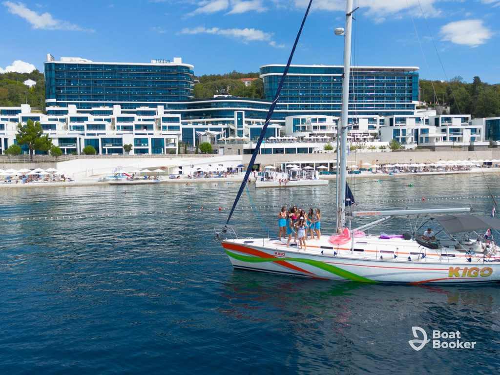 A group’s sailing in Croatia on a sailboat near a modern beachfront hotel, with a beach and umbrellas lining the scenic shore.