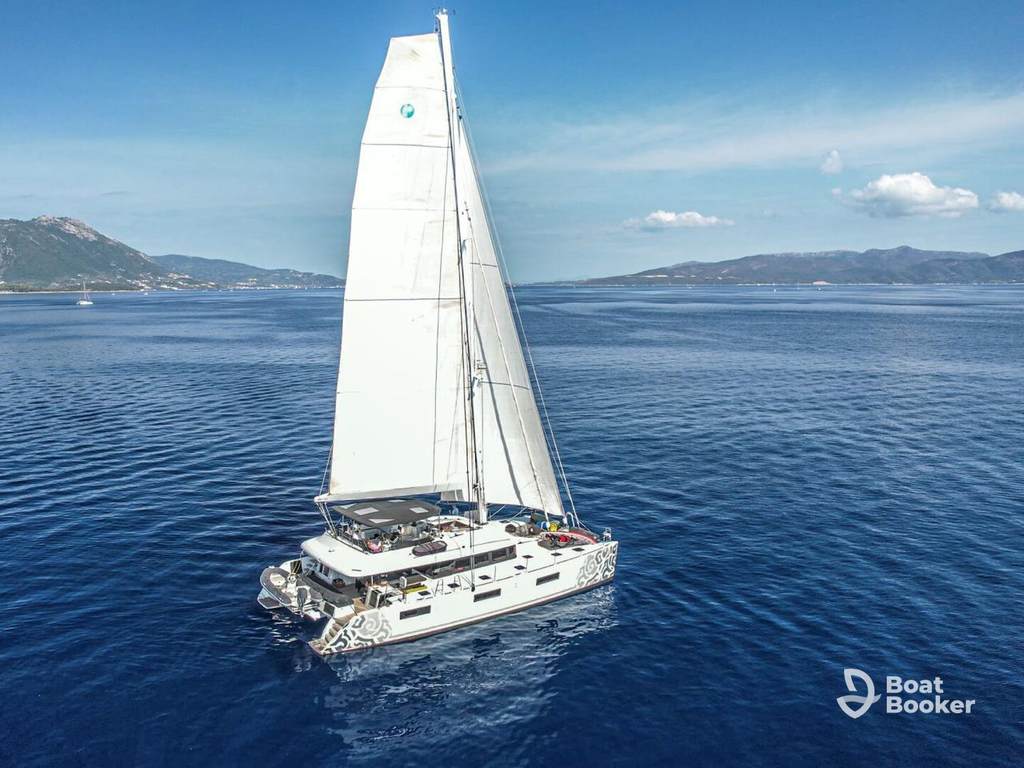 A white catamaran with raised sails glides across calm blue water near a mountainous coastline—it’s perfect for sailing in Croatia. The BoatBooker logo’s in the lower right corner.