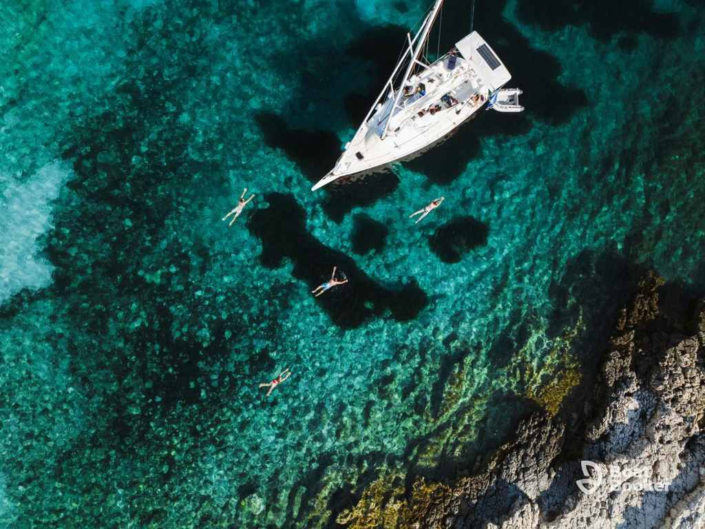 An aerial view of a sailboat anchored by rocky shore, with several people swimming in clear turquoise water—it’s perfect for sailing Croatia. No species like Tuna or Grouper are visible here.