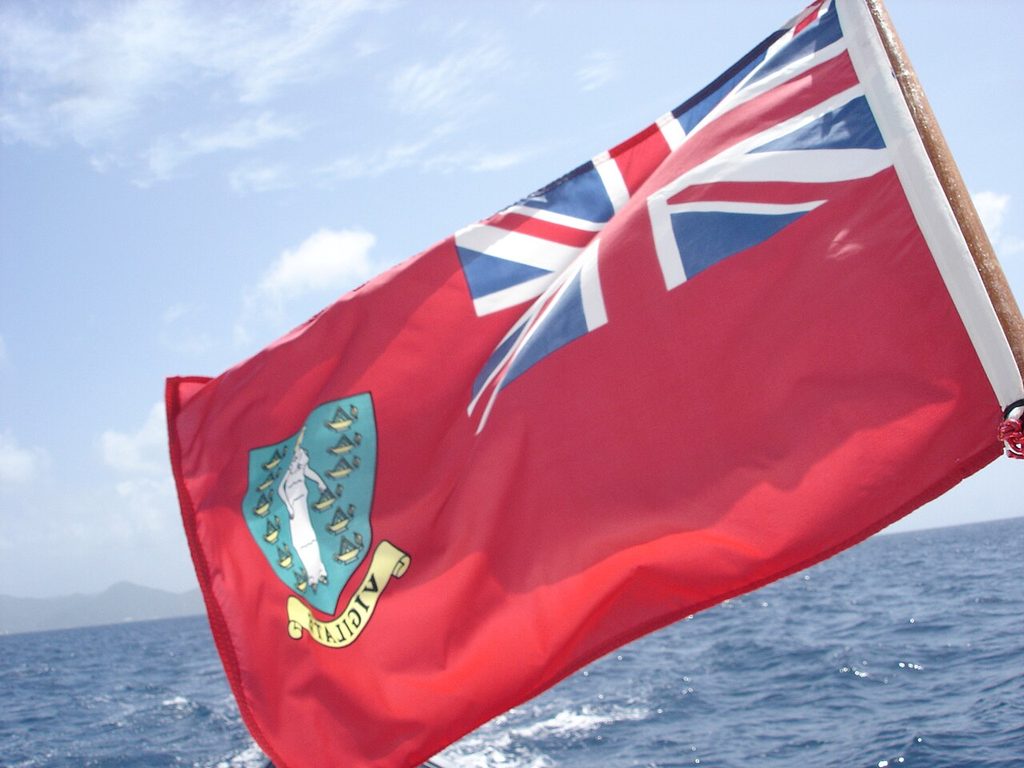 A red ensign flag with the Union Jack and a crest showing a woman and lamps flutters on a boat above a blue ocean under a clear sky.