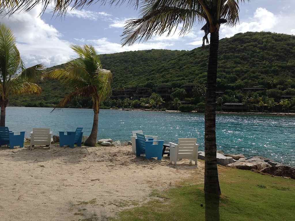 Chairs and tables are set on sandy ground by a blue body of water, with palm trees and green hills behind under a partly cloudy sky.
