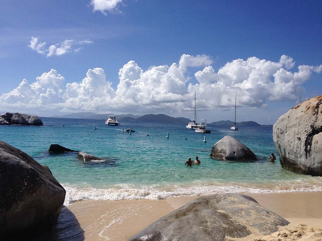 There’s a beach scene with large rocks, clear blue water, swimmers, and sailboats anchored offshore under a partly cloudy sky.