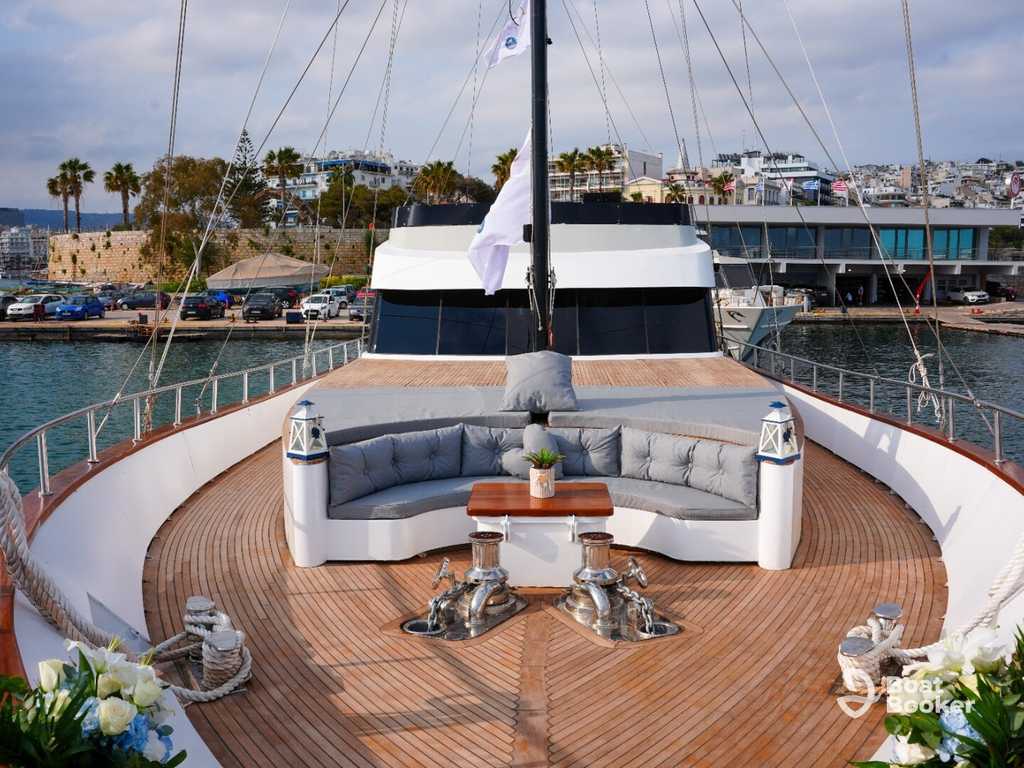 The yacht’s deck has a curved seating area, cushions, a small table, and two metal anchors. It’s docked in a marina with buildings and Palm Trees in the background.