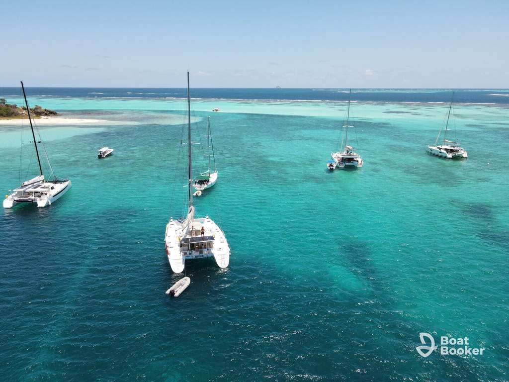 Five catamarans are anchored in clear turquoise water by a small island, with a visible reef and blue sky. The Boat Booker logo’s in the bottom right corner. No fish or species are shown.