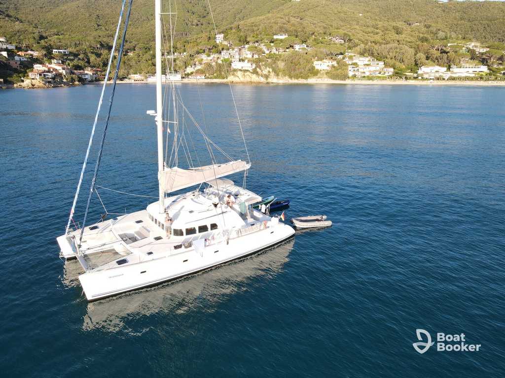 A white catamaran yacht’s anchored on calm blue water near a coastal area with green hills and houses in the background. BoatBooker logo’s in the corner.