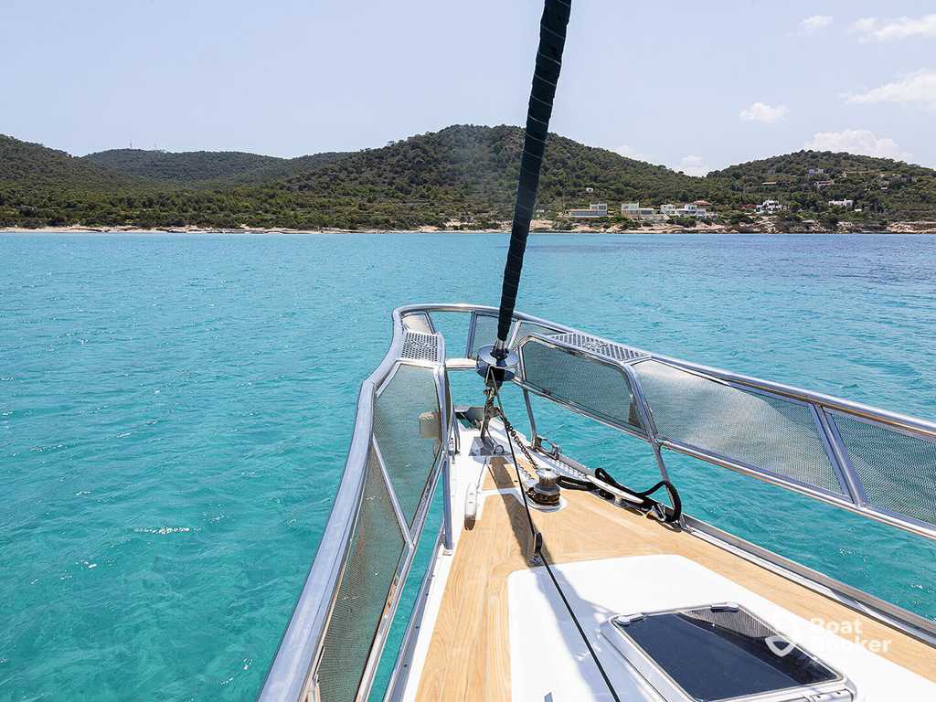 You’re looking from the bow of a yacht on clear turquoise water, sailing toward a coast dotted with hills, trees, and some buildings beneath a sunny sky.
