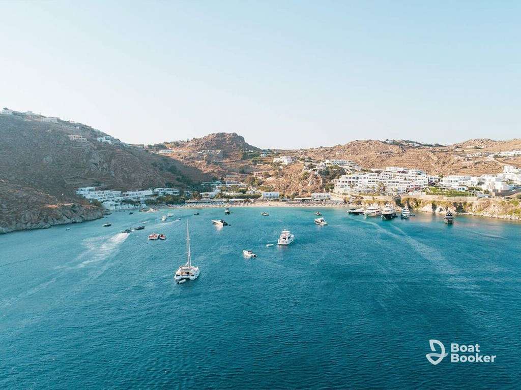 Several boats and yachts are anchored in a calm bay surrounded by rocky hills and white buildings under a clear sky. The Boat Booker logo’s in the lower right corner.