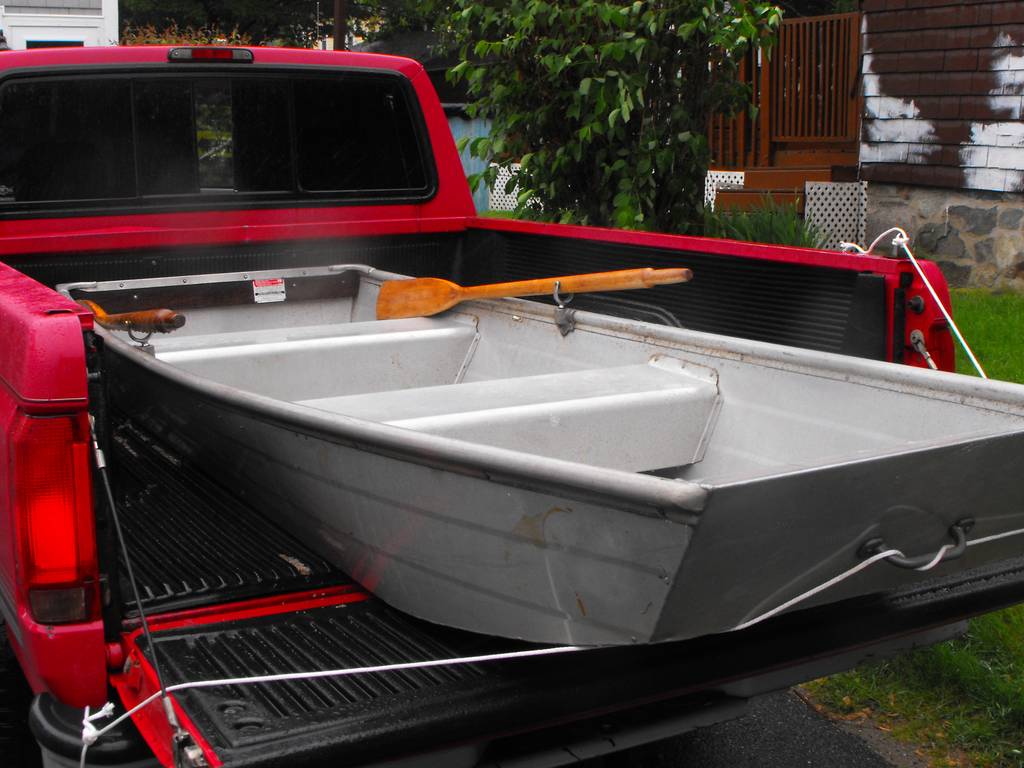 A small aluminum rowboat with wooden oars’s loaded into the bed of a red pickup truck, parked in a residential driveway.