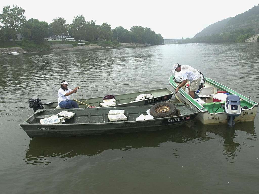 Two people in small boats are collecting debris, like a tire, from the river. Trees and houses line the riverbank behind them.