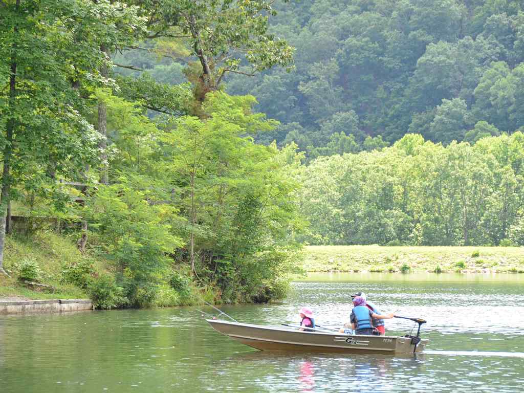 Two people in a small motorboat are traveling on a calm lake that’s surrounded by green trees and forested hills on a bright day.